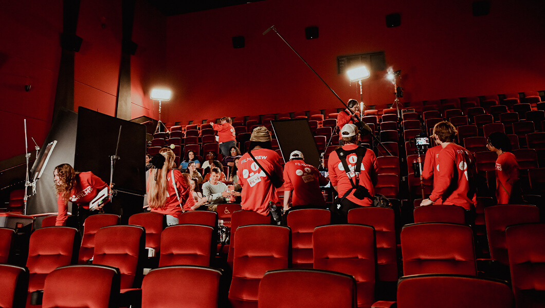 Film crew in Coca-Cola Refreshing Films shirts set up lights and gear inside a theater with rows of bright red seats.