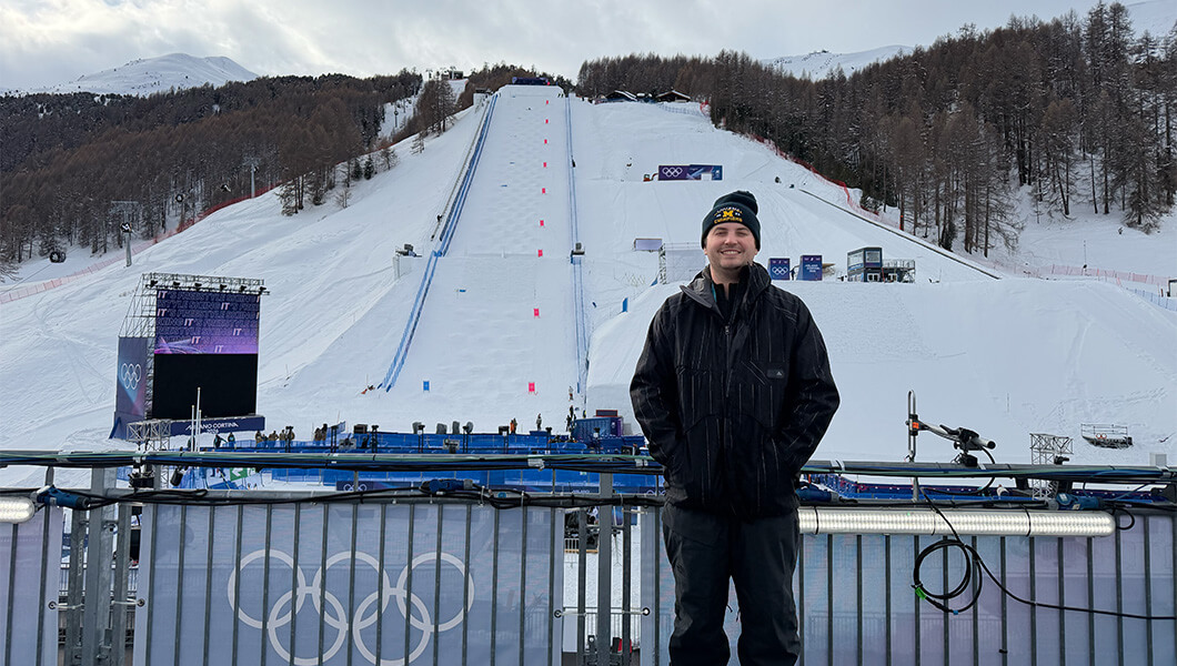 Cameron Champagne stands in front of a large, snowy hill with trees and mountains in the background. He is wearing a black winter coat and beanie.