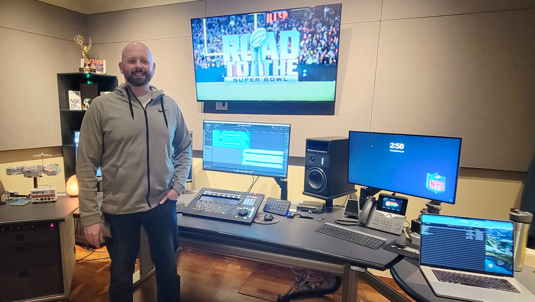 Tom McWilliams stands in a room with a few desks with computer screens and speakers. A large screen on the wall behind him displays “Road to the Super Bowl” over a shot of a field, goalpost, and fans in the stands.