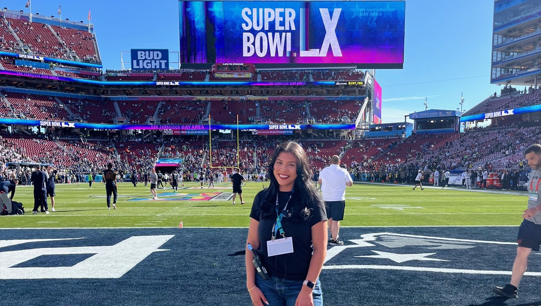 Michelle Navarrete smiles at the camera and stands on a football field with crowded stands behind her and crew members around her. A large screen behind her displays “Super Bowl LX” with a blue and pink background.
