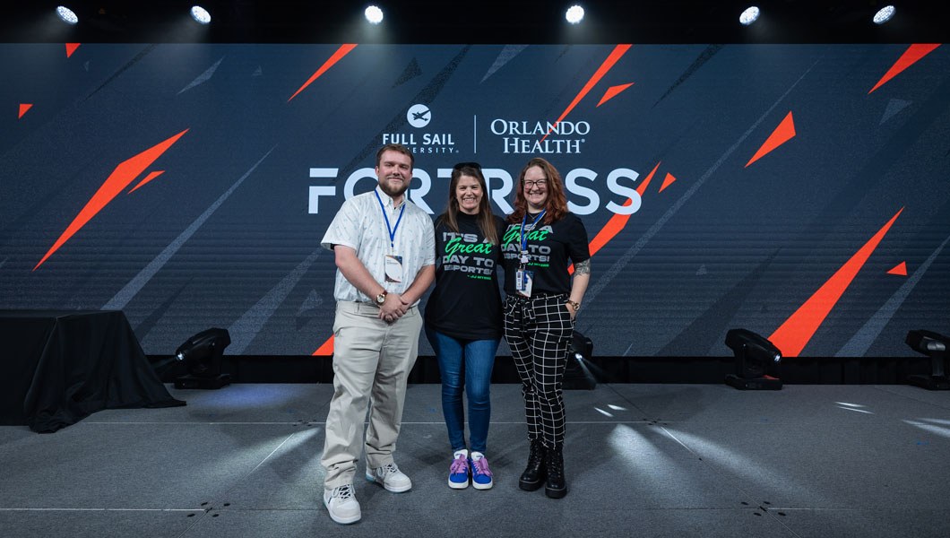 A man and two women stand together and smile. The words Full Sail University Orlando Health Fortress are on the wall behind them.