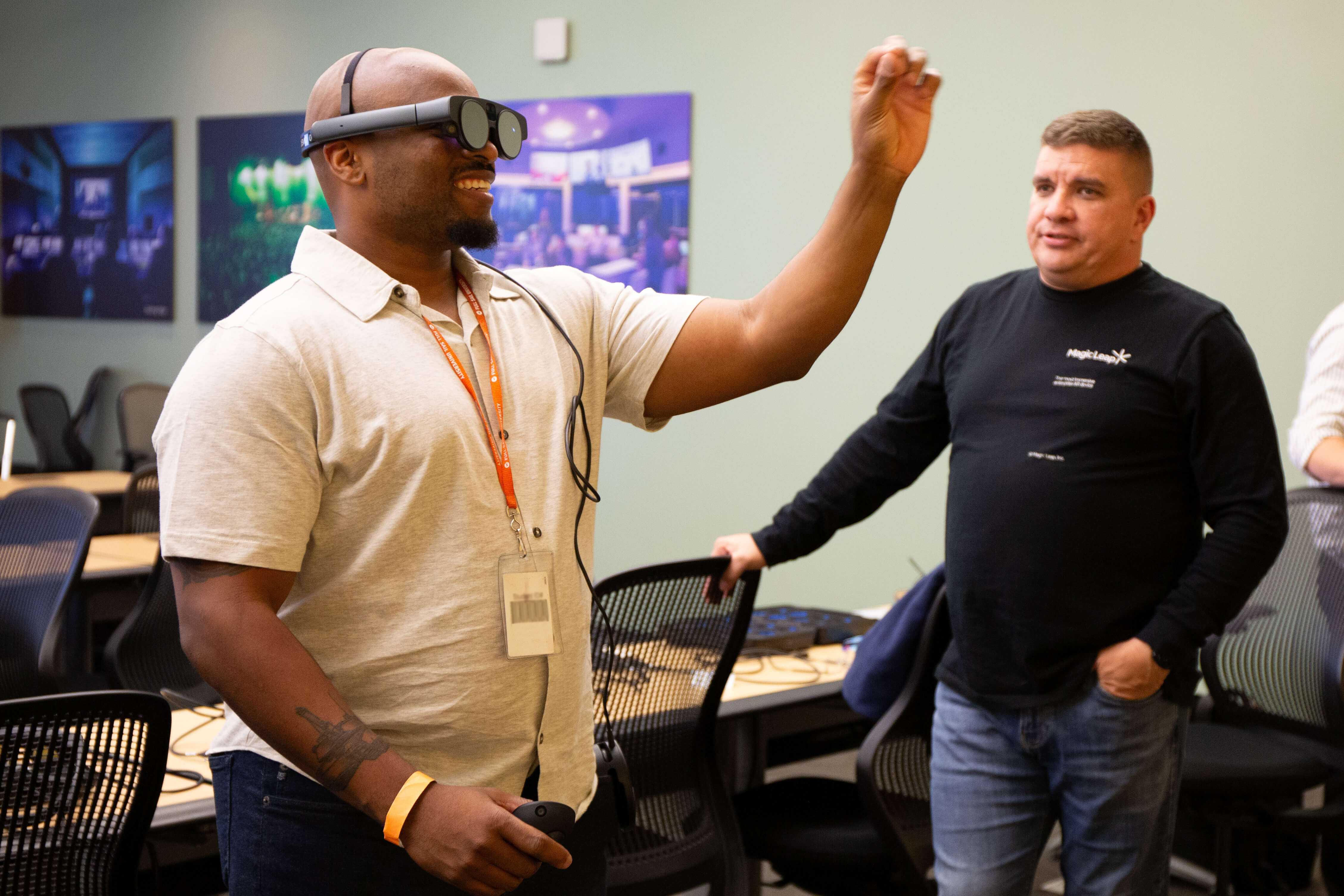 A Full Sail student wears a VR headset, a hand raised to interact with what they see, and a person stands to the side, observing.