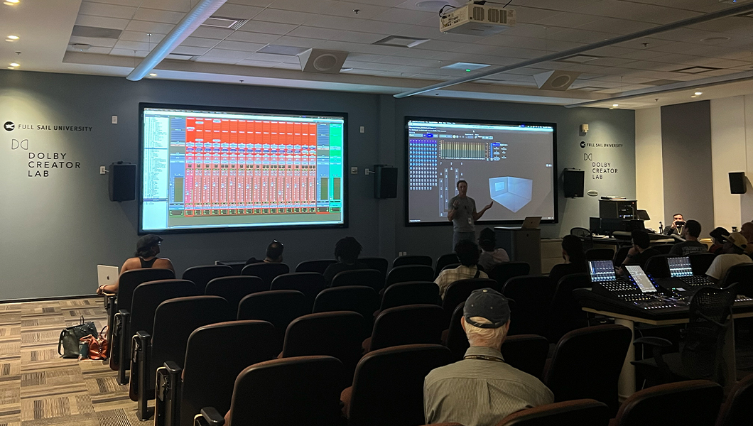 Darren Schneider stands at the front of a room and speaks to an audience. Two projector screens are behind him displaying Pro Tools software.