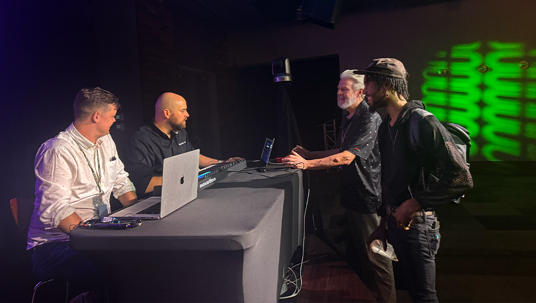 Scott Dickinson and Evan Weiss sit at a table with laptops and a keyboard. Two audience members stand by the table and talk with them.