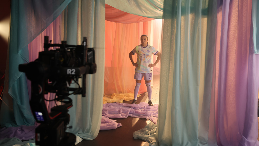 An Orlando Pride player poses for the camera with their hands on their hips. They are wearing a pastel uniform and surrounded by colorful fabric draped around the set.