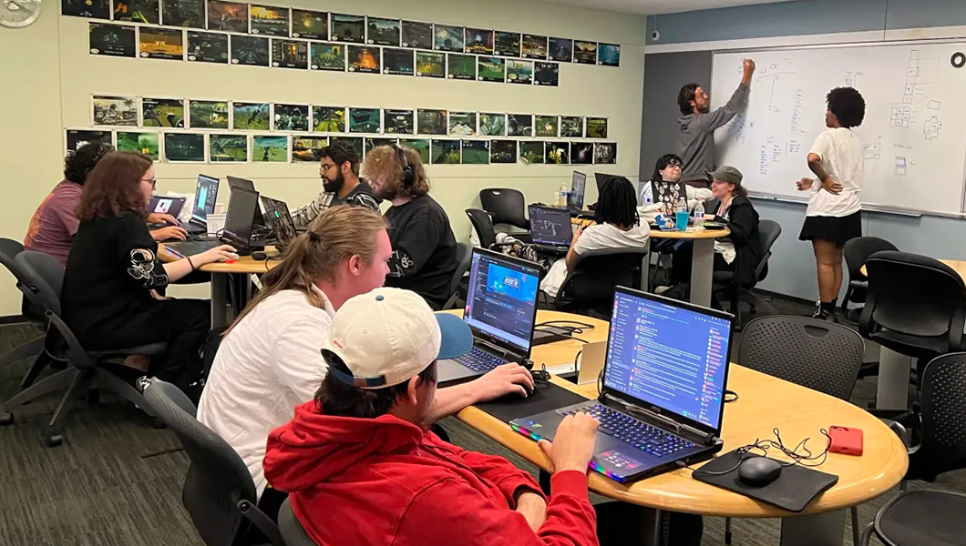 Several students sit at desks together on their laptops. One student stands and writes on a whiteboard. The back wall is covered in rows of images from video games.