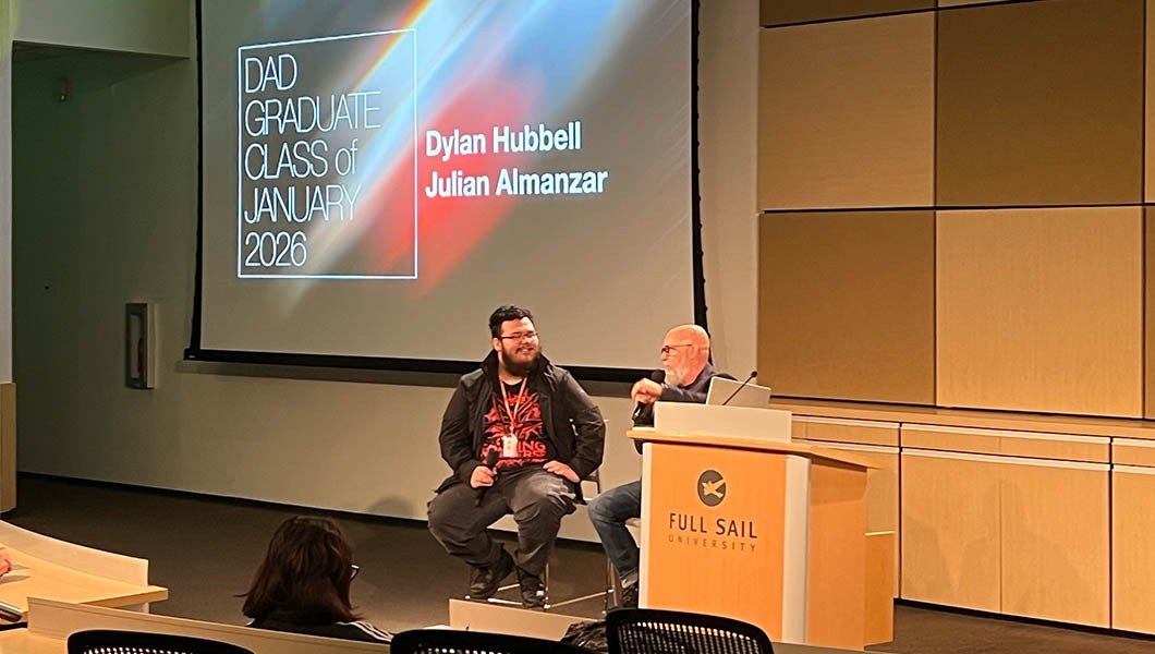 Dylan Hubbell and Joel Sink sit in front of an audience and speak into microphones. A screen behind them displays “DAD Graduate Class of January 2026.”