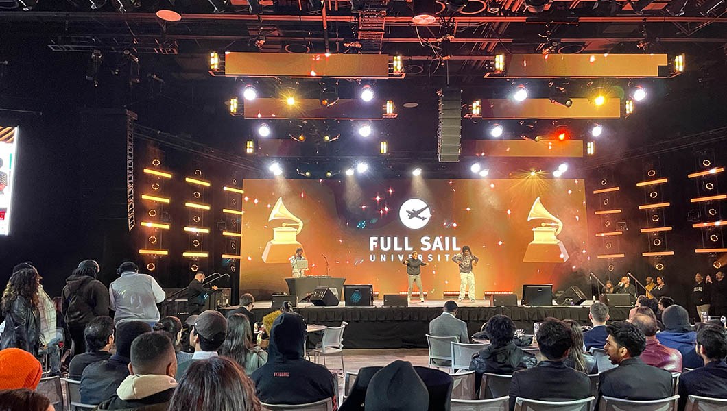 An audience of students sit in chairs facing a stage with a live DJ and emcees. A large screen behind them displays “Full Sail University” with two Grammy Awards.