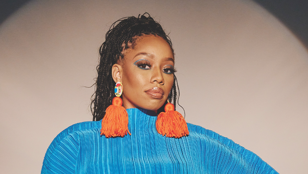 DOE stands with her hand on her hip. She has long braids tied up in a ponytail and wears a bright blue sweater and pants and earrings with orange tassels.