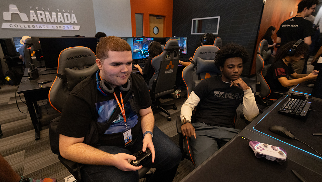 A student sits at a gaming console holding a controller and wearing headphones around their neck while another student sits nearby and watches. Other students play games around the room at various consoles and a sign on the wall displays “Full Sail Armada Collegiate Esports.”
