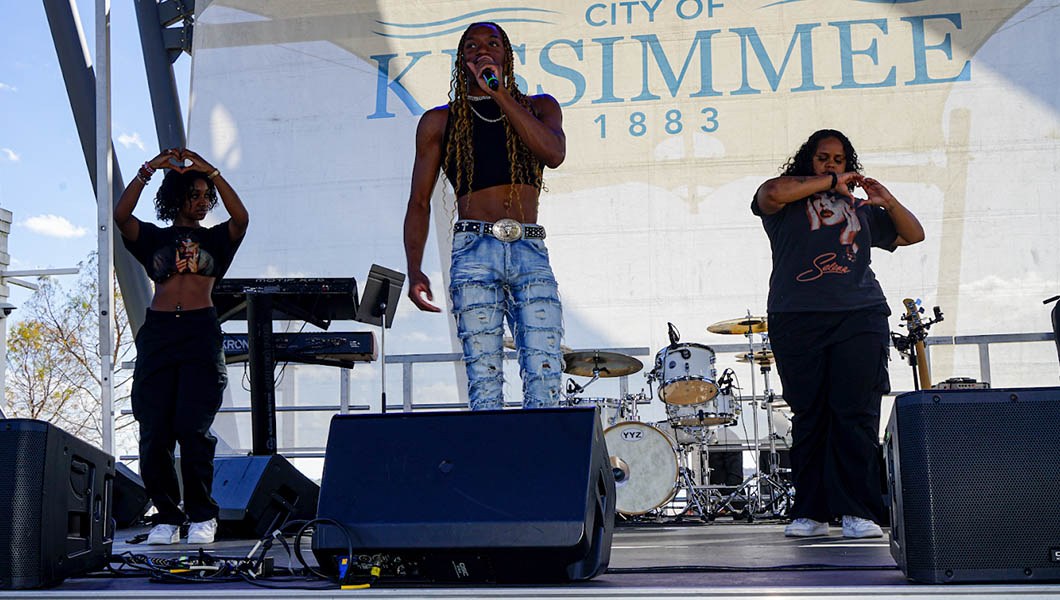 Three students stand on a stage. One sings into a microphone and two form their hands into hearts. A banner behind them reads “City of Kissimmee 1883.”