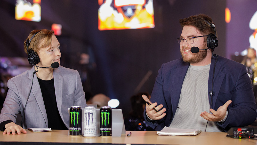 Two well-dressed people are seated at a hosting desk while wearing headsets and chatting.