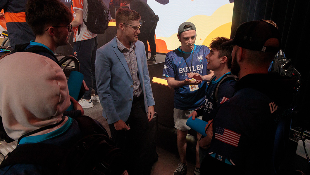 A man in a light blue sport coat stands near a stage while talking to a group wearing Butler Esports jerseys.