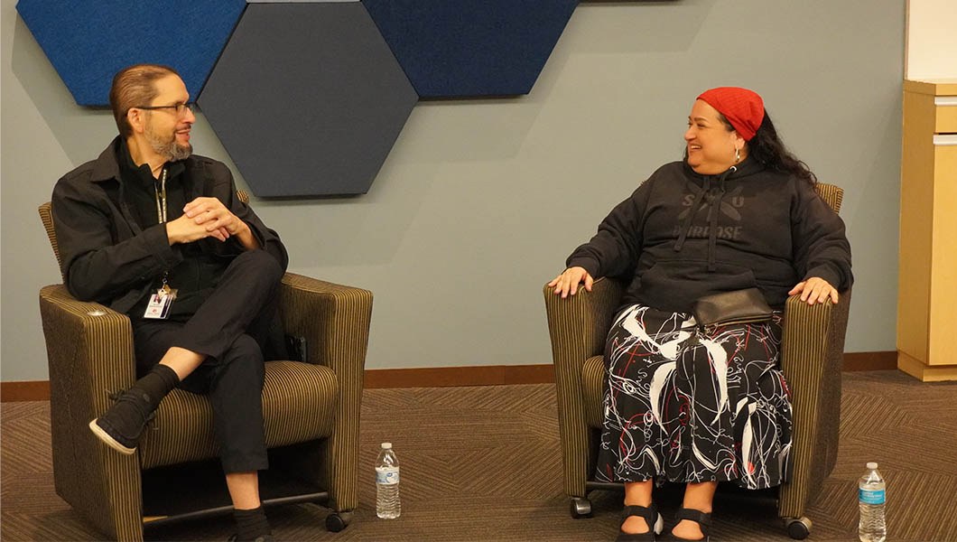 Israel Vasquetelle and Nicole Sanzio sit in chairs and talk to each other. Israel wears a black jacket and pants, and Nicole wears a black hoodie, patterned pants, and a red bandana.