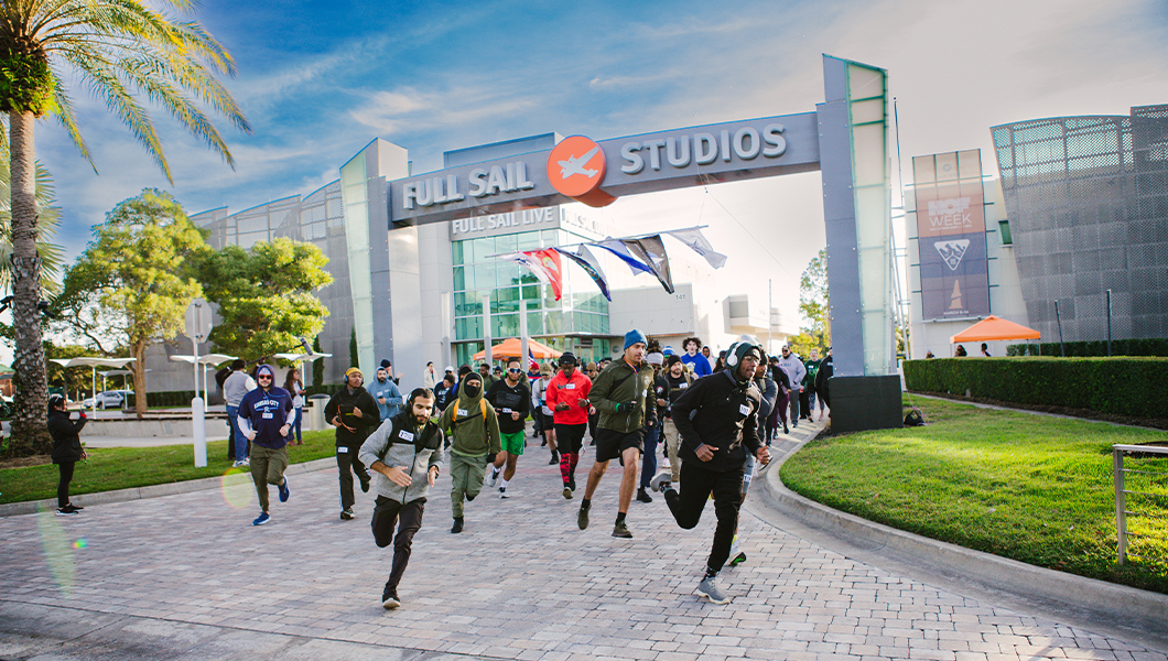 A crowd of people run under the Full Sail Studios archway wearing athletic gear. On either side of the archway are trees and green grass with buildings behind it.