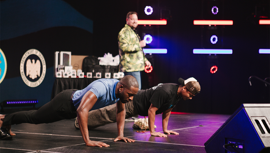 Two veterans do push ups on a stage with red and blue lights shining. Aaron Hall stands behind them watching and wearing a camo green blazer.