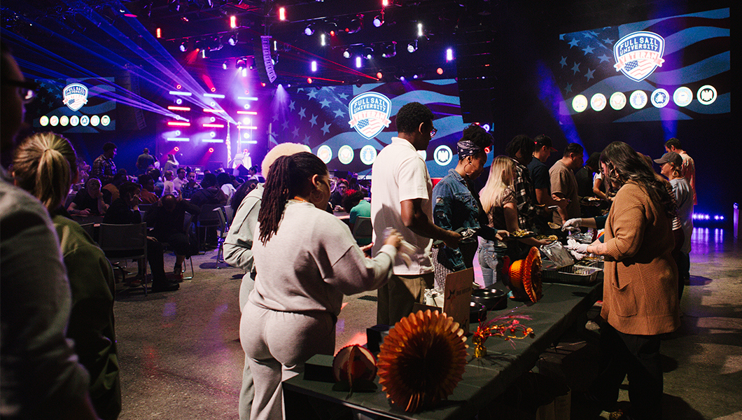 People stand in a line to get food from a long table with various Thanksgiving decorations on it. People on the other side of the table serve them food. Large screens display the Full Sail University Veteran logo while red and blue lights shine.