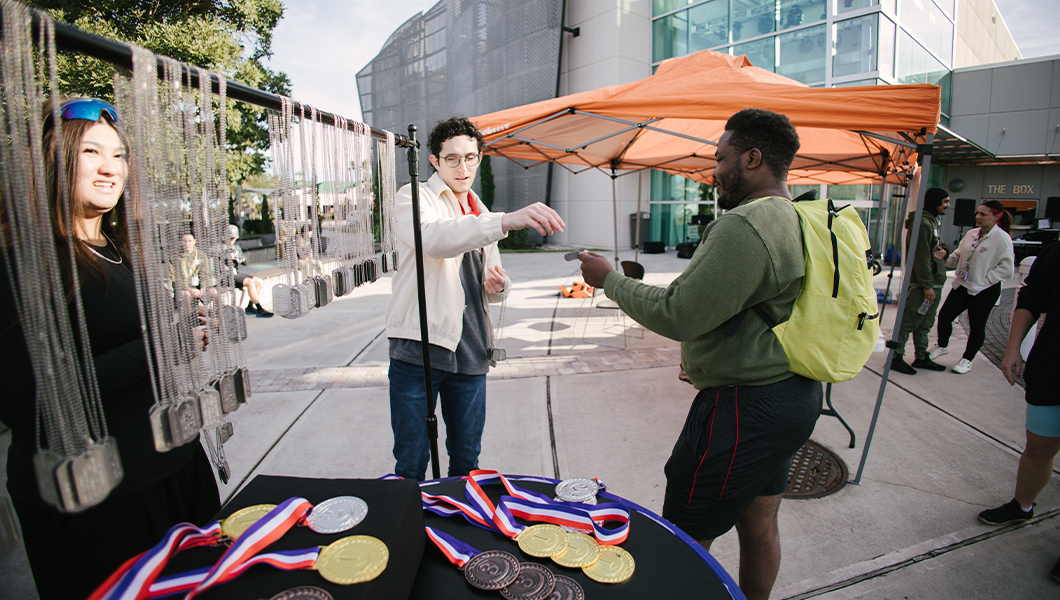 Two people stand next to a table with gold, silver, and bronze medals arranged on it and a rack of silver dog tags hanging above it. One person hands a dog tag to a veteran.