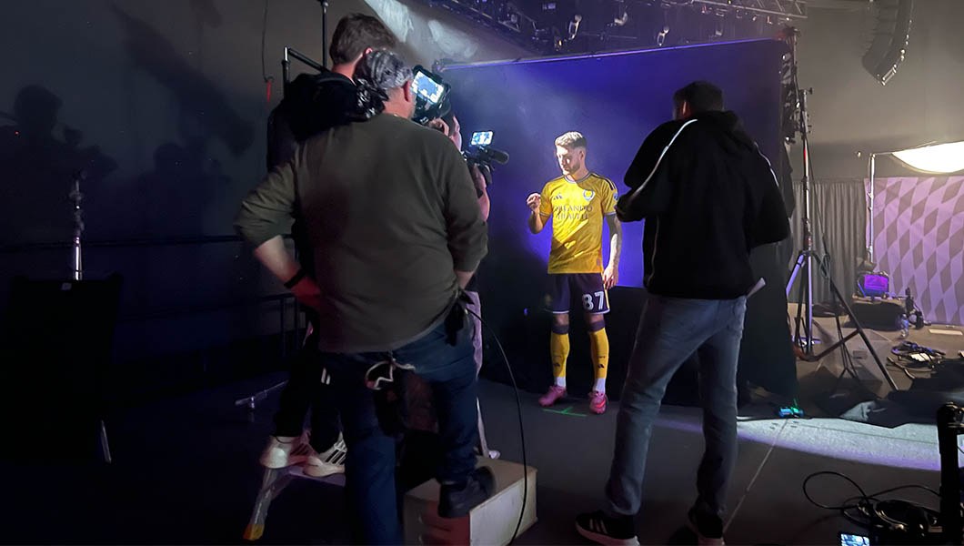 An Orlando City player wears a yellow and purple uniform and poses for the camera in front of a purple backdrop while production crew stand around and capture the shot.
