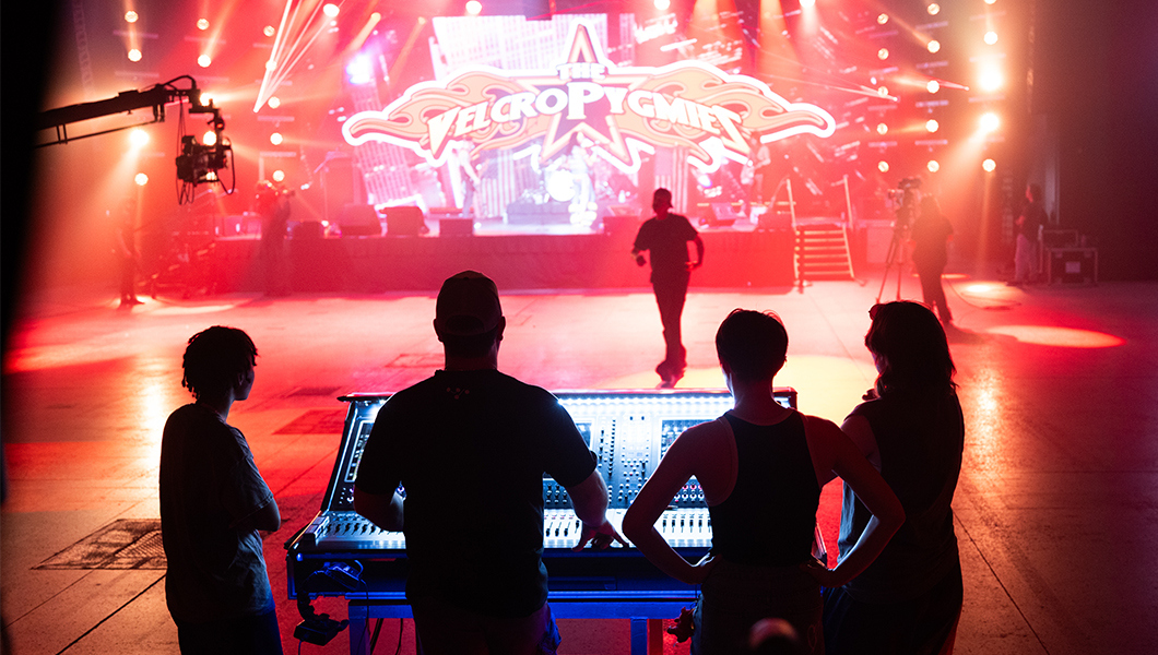 Students stand around a soundboard with a production crew member showing them how to use it. A drum set is on the stage in front of them with a screen displaying “Velcro Pygmies” and bright orange and red lights.