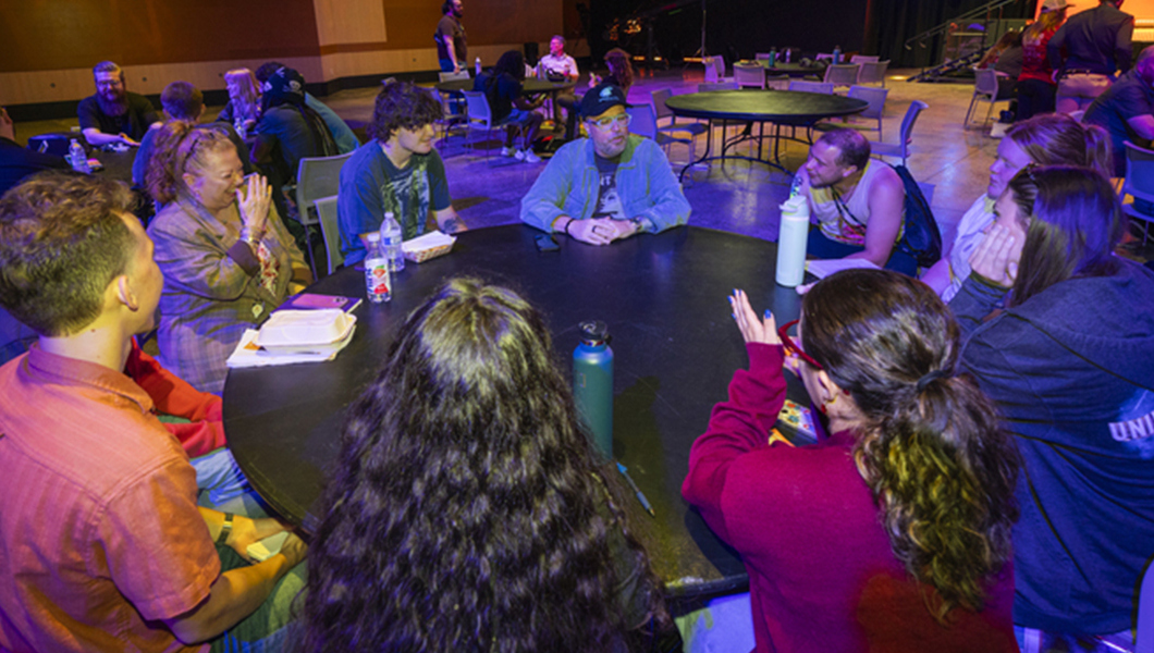 Several students sit around a round table with Aaron B. Koontz. Other round tables are around the room with students talking to other grads.