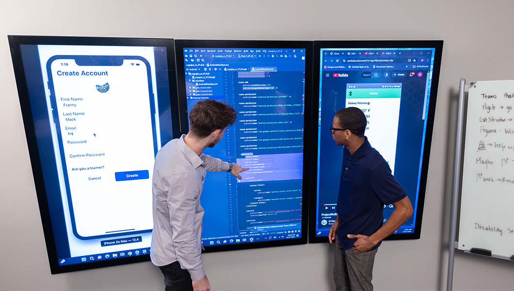 Students review app code and interface layouts on wall-mounted touchscreens in the Smart Lab.