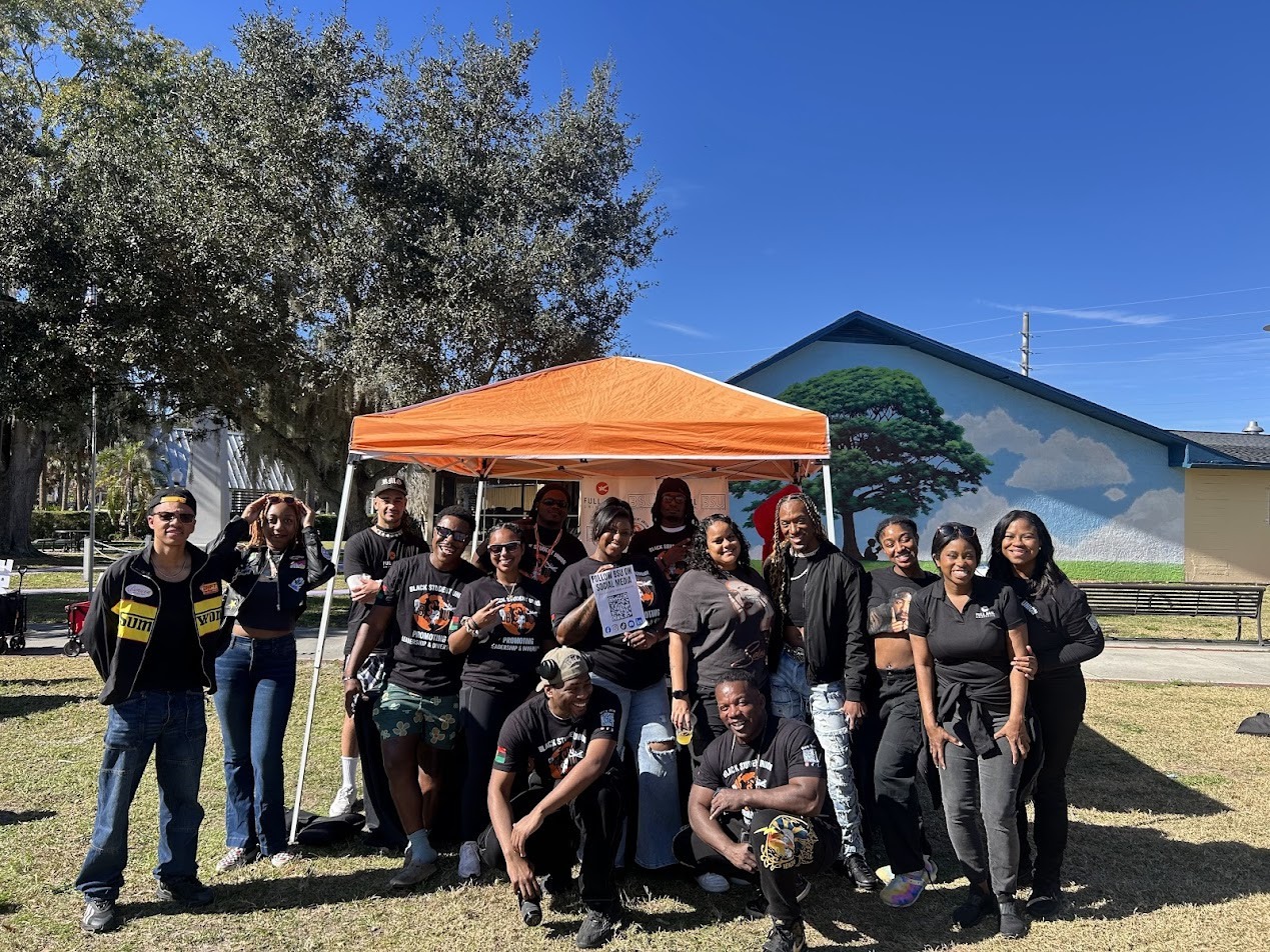 A group of staff and students stand under an orange tent and smile at the camera. Several of them are wearing matching black t-shirts with the Black Student Union logo.