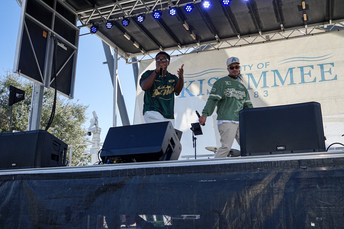 wo students stand on a stage wearing green jerseys and sing into microphones. There are speakers in front of them on the stage and a banner behind them displays “City of Kissimmee.”