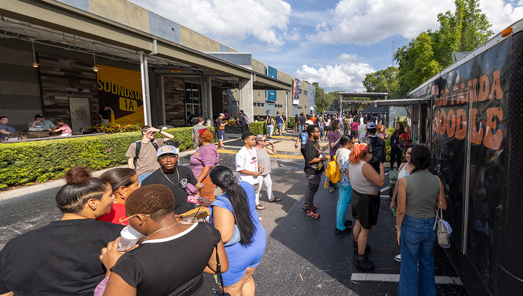 Crowds of attendees line up at food trucks on a blacktop with blue skies above and a stage in the distance.