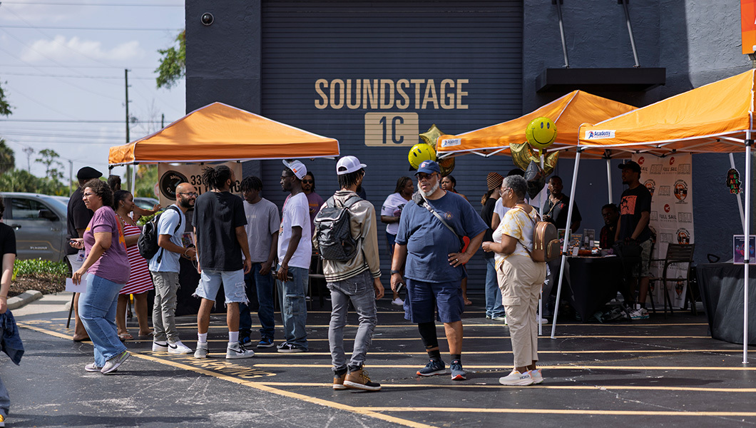 Groups of attendees standing on a blacktop beside orange tents, a garage door on the wall reads ‘Soundstage 1C.’