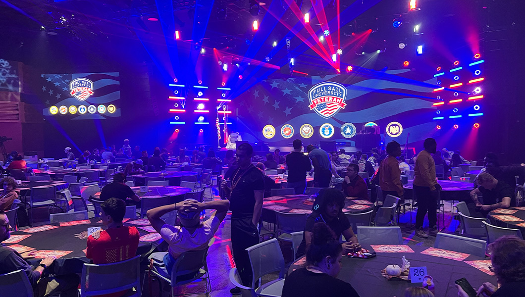 People gather at many round tables in a large room with a stage. Two screens display the Full Sail University Veteran logo as well as logos of military branches. Red and blue lights shine brightly.