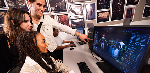 A student sits at a computer displaying 3D images while two other students stand next to them and one points to the screen. Another student is behind them sitting at a computer. Pictures of 3D renderings hang on the wall behind them.
