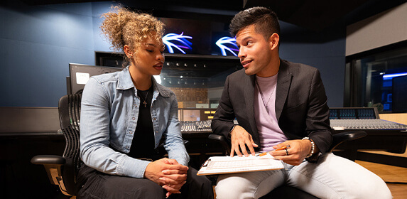 Two students sit next to each other. One holds a clipboard and points to it with a pen while the other looks at the clipboard. A sound board and multiple screens are behind them.