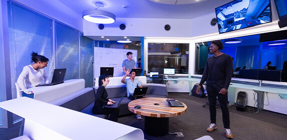 Five students gather in a large room with windows and two TV screens. Four of them sit with laptops and one stands facing them with a laptop on a nearby table.
