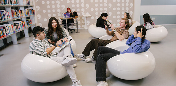 Several students sit in white bubble chairs in a large room with a wall of bookshelves on either side. One student sits at a table in the back corner with a laptop and a bag.