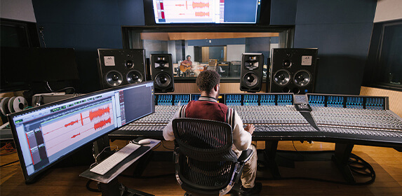 A student sits at a sound board surrounded by speakers and a large monitor. On the other side of a window in front of them another student plays a guitar.