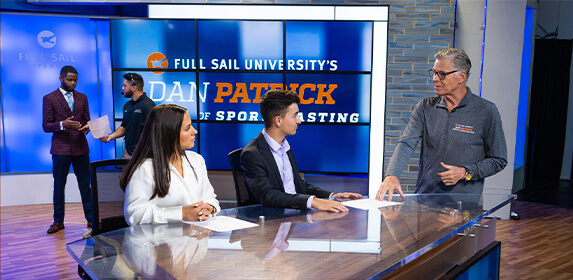 Dan Patrick speaks to two students sitting at a table. Behind them, a large screen displays “Full Sail University’s Dan Patrick School of Sportscasting.” Two other students stand nearby, one wearing a suit and one wearing a headset and polo shirt and holding a piece of paper.