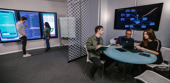 Two students stand in front of a large screen mounted on a wall that displays a mobile phone screen. Behind them, three students sit at a round table with a laptop.