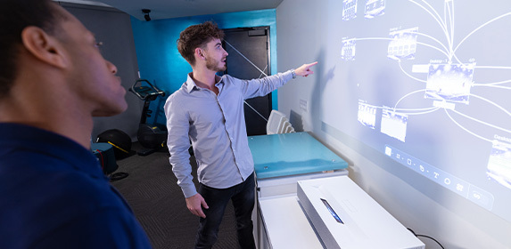 Two students stand in a classroom and look at a projection of multiple computer screens on the wall.
