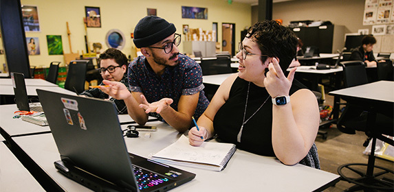 Two students are seated in a computer animation classroom in front of their laptops while an instructor stands between them mid-discussion.