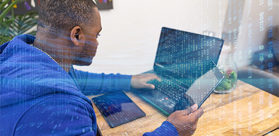An overlay of code appears on an image of a computer science student while they work concurrently on their laptop and tablet.