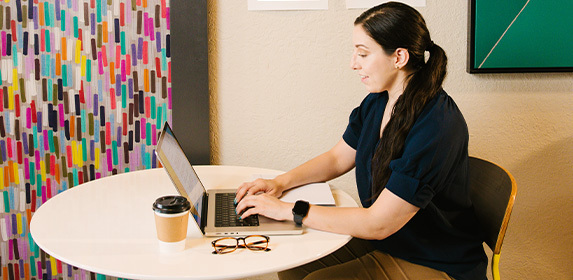 A student wearing a blue sweater sits at a table. They are typing on a laptop and a coffee cup and eyeglasses are on the table next to them.