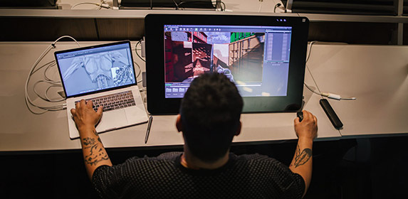 A digital arts & design student is seated at their workstation which features a MacBook and a large tablet where the 3D model from their laptop is rendered on the alternate screen.