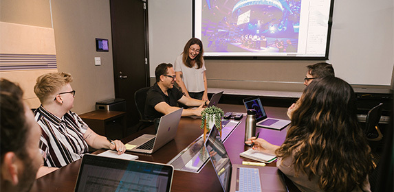 A group of five people are seated at a table in a conference room while a woman in the front of the room looks on smiling and pointing at a seated attendee's laptop.