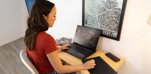 A Game Art student sits at a desk. They are using a tablet to draw a 3D-rendered face, which is displayed on their laptop.
