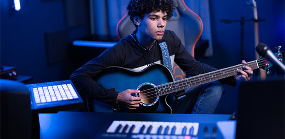 A music production student is playing an acoustic-electric bass guitar while seated in front of a MIDI keyboard and launchpad from their Project LaunchBox™.
