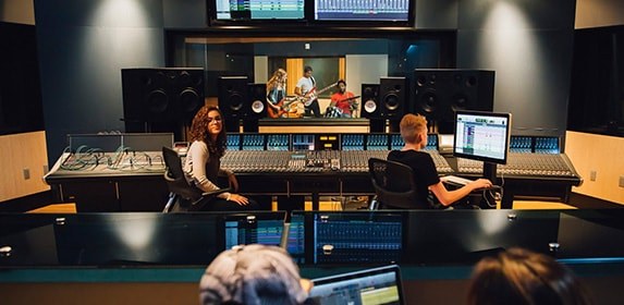 A group of students monitors audio levels on a massive soundboard for a band playing in a recording studio classroom.