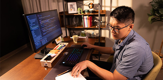 A web development student consults his notepad while writing code on his desktop computer in a home office.