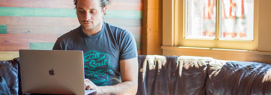 A student in a blue Full Sail t-shirt sits on a leather sofa and types on a laptop.