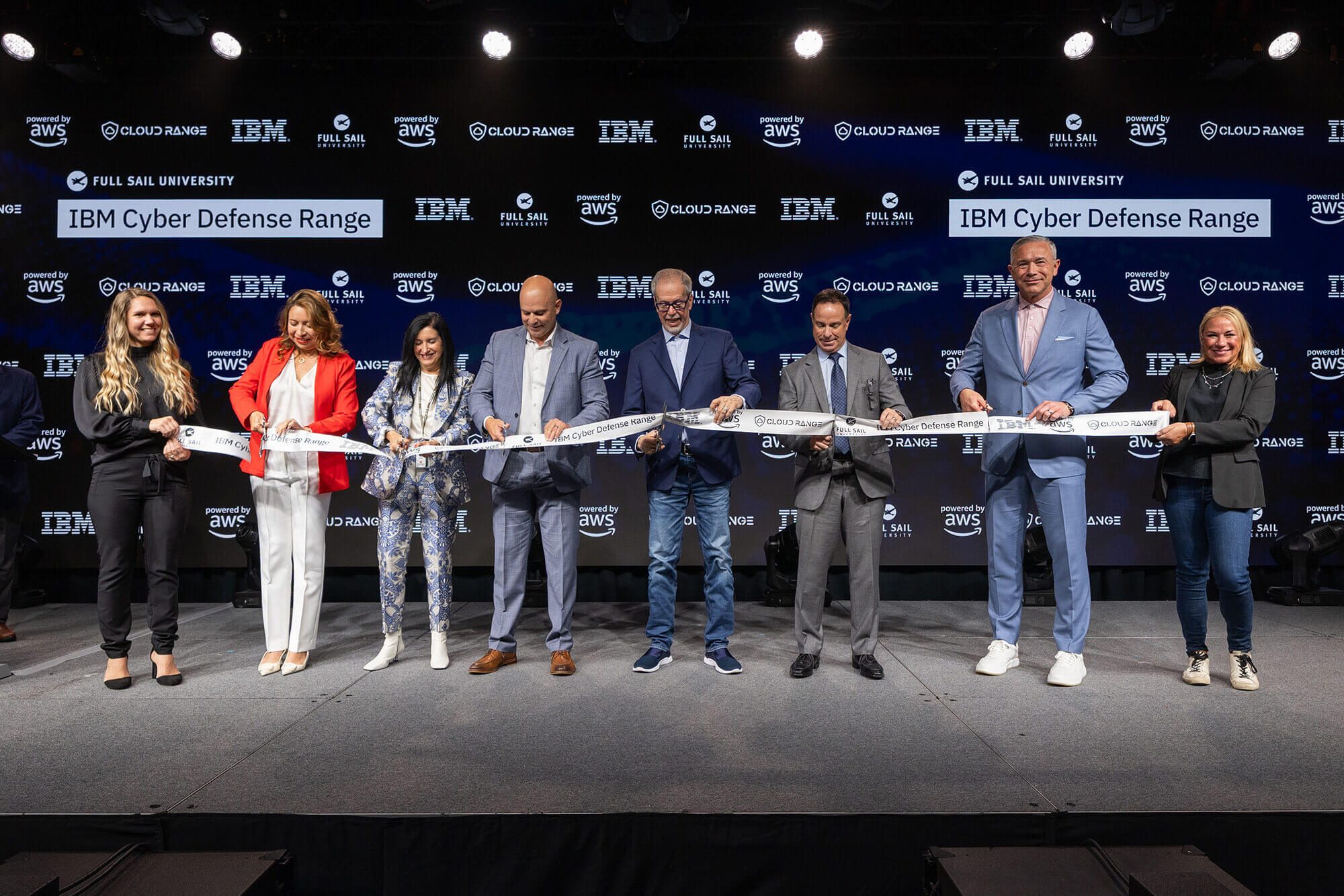 Eight people each hold scissors and cut into a long white ribbon in front of a screen saying Full Sail University IBM Cyber Defense Range.